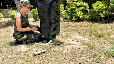 Camo-clad woman handling mummified slave legs in BDSM outdoor session