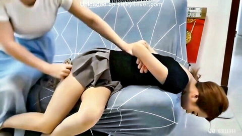 East Asian brunette spreads legs wide as friend fingers her pussy under gray skirt on couch