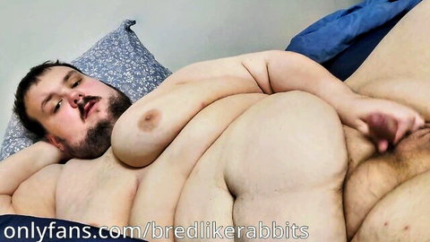 Obese gay bear lying nude on bed with massive belly rolls and moobs exposed