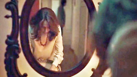 Brunette woman sitting clothed in front of ornate mirror in dimly lit room