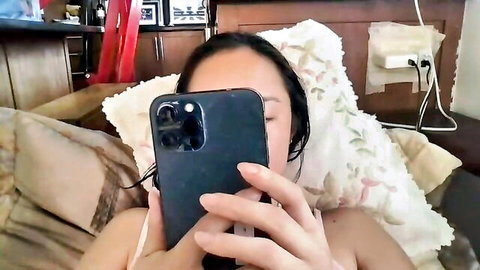 Asian babe in tank top taking flirty selfie on bed pillows teasing cleavage