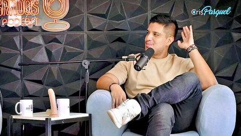 Podcast host gesturing excitedly next to realistic dildo on table in Jugs Podcast studio