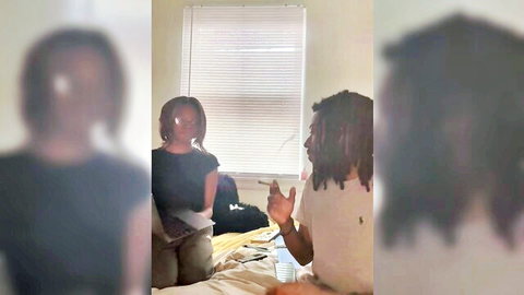 Blurry ebony couple sitting clothed on bed chatting in amateur bedroom