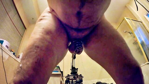 Hairy chubby man in metal chastity cage stands legs spread over fucking machine dildo aimed at his ass