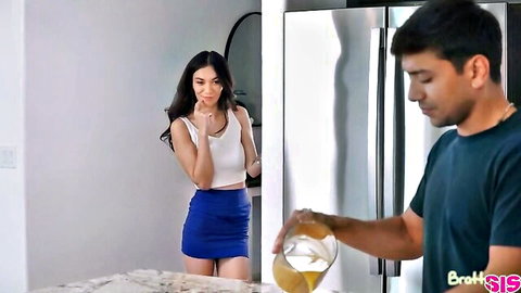 Skinny brunette teen in tight white tank top and blue mini skirt curiously watches guy pour thick white liquid from jar in kitchen