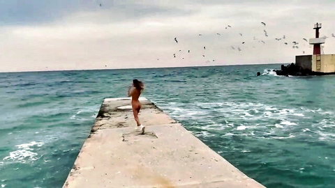 Nude slim woman walks on Sochi beach pier by sea and lighthouse