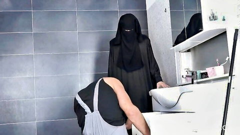 Veiled Arab niqab woman watches bent-over maid scrubbing sink in tiled bathroom