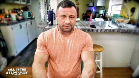 Muscular man in orange shirt sits in kitchen with All in the Family watermark