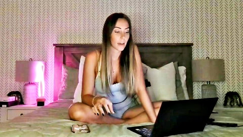 Petite brunette in tight blue lingerie sits cross-legged on bed using laptop webcam