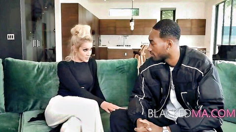 Blonde MILF in tight white pants sits close to black stud on couch flirting intimately.