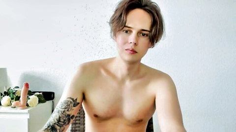 Tattooed shirtless twink sits nude with huge pink dildo nearby on webcam