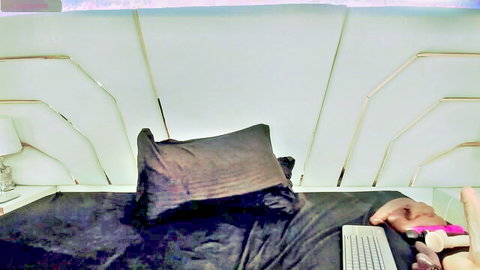 Hands gripping thick pink dildo on dark bed sheets next to keyboard in amateur bedroom