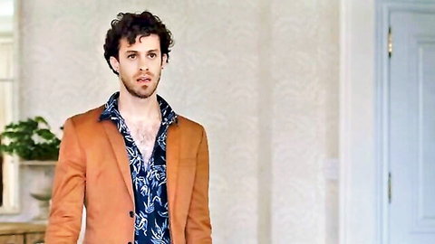 Curly-haired man in open orange blazer and shirt exposing chest in luxurious room