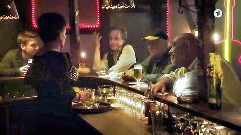 Sexy brunette in black top chats with group of clothed men at neon-lit bar counter.
