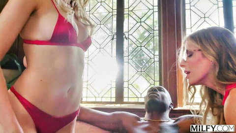 Two long-haired blondes in skimpy red bikinis tease muscular black man lying down in interracial threesome foreplay