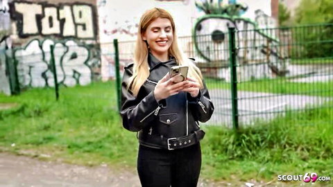 Smiling blonde babe in sexy leather jacket holds phone outdoors by graffiti fence Scout69