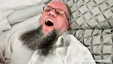 Elderly bearded man in white shirt lying on bed, mouth open in ecstasy