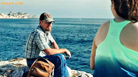 Man in plaid shirt and jeans sits on rocks watching curvy woman's back in green tank top by ocean sea
