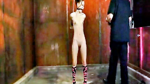 Naked ballgagged woman suspended by wrists in dungeon wearing striped stockings with suited man behind