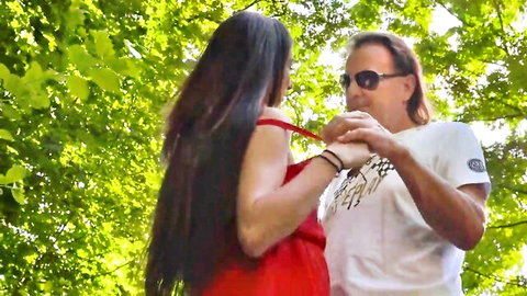 Young woman in red dress holds hands intimately with older long-haired man outdoors