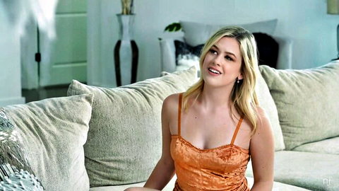 Blonde babe in tight orange velvet top smiling with cleavage tease on couch