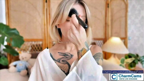 BambiFawn blonde cam girl applying makeup in open white robe showing tattoos