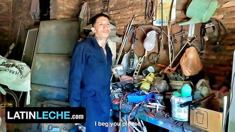 Tattooed muscular Latino mechanic in open blue jumpsuit poses seductively in cluttered workshop