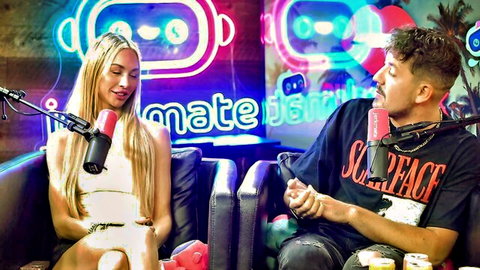 Blonde woman in white top smiles while chatting with curly-haired guy in Scarface shirt on neon-lit podcast couch