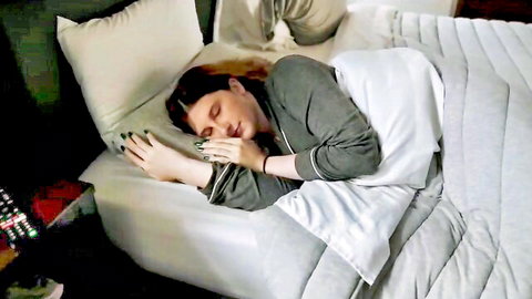 Brunette woman sleeping in gray pajamas on white bed sheets in dim bedroom.