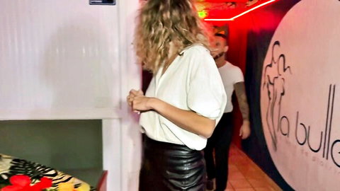Blonde woman in open white blouse showing cleavage teases tattooed guy in leather skirt at neon club