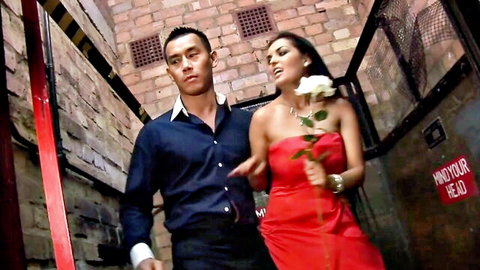Curvy brunette in red dress holding rose walks with boyfriend in brick alley foreplay
