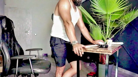 Muscular bearded hunk in tight white tank top and shorts posing sensually by table