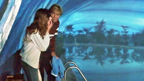 Brunette woman in white top and jeans chats with blonde hunk by indoor pool