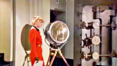 Blonde woman in red uniform and gloves stands profile by vintage spotlight.
