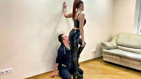 Femdom mistress in latex pants stands tall as kneeling slave presses face into her shiny latex ass