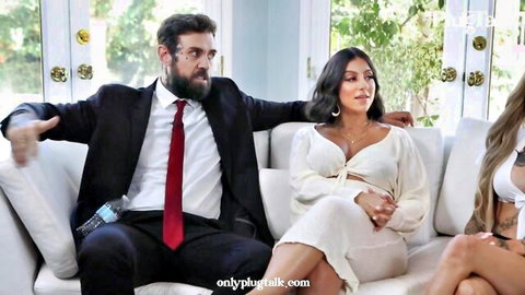 Bearded man in suit gestures between curvy brunette and blonde women on couch showing cleavage