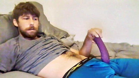 Bearded hairy guy lying on bed gripping huge purple dildo over his blue track pants bulge
