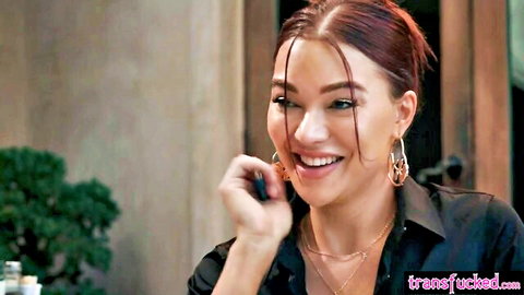 Leana Lovings smiling seductively in black blouse, red hair, hoop earrings