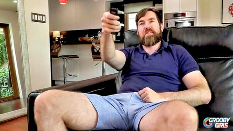 Bearded man in shorts relaxes on couch, GroobyGirls Genesis Green scene