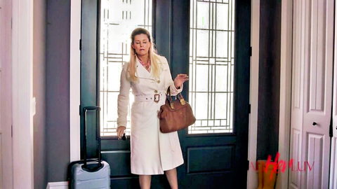 Blonde MILF in tight white trench coat stands with brown purse and rolling suitcase at door