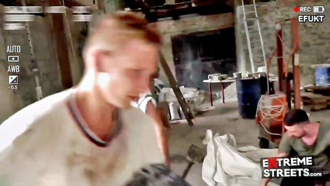 Two sweaty men wrestle roughly on white sheet in gritty warehouse