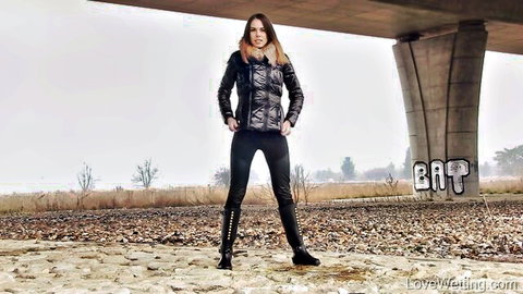 Jessica Bell posing in black fur-lined jacket, tight pants and boots under bridge in wetting fetish scene