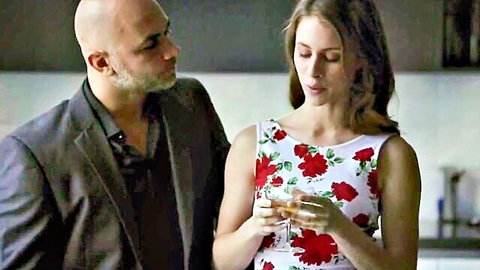 Brunette babe in floral rose dress holds champagne glass flirting with bald suited daddy in kitchen