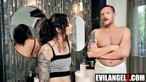 Tattooed curvy woman in checkered crop top smirks at shirtless muscular man in towel in bathroom mirror.