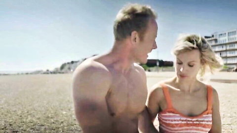 Blonde MILF May Kelly embracing shirtless hunk on windy beach