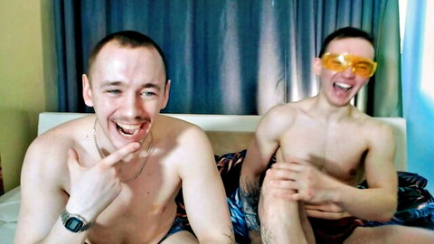 Two shirtless twinks in colorful swim trunks laughing on bed with orange sunglasses