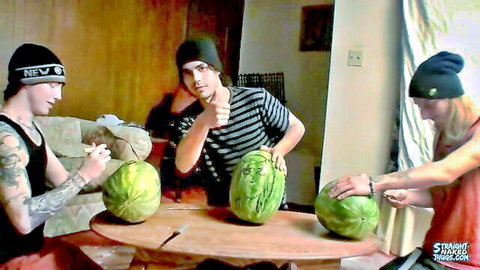 Three tattooed young men in beanies signing huge watermelons at a wooden table indoors