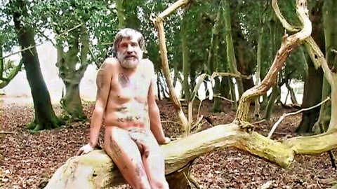 Mature bearded nudist covered in white paint sitting naked on log in forest