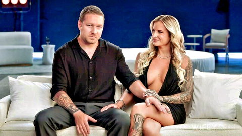 Tattooed blonde wife in plunging black dress flashes deep cleavage sitting with tattooed husband on couch.