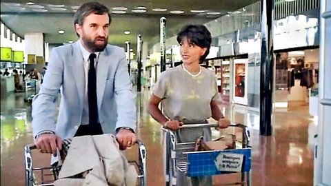 Bearded man in suit and petite short-haired woman in dress pushing shopping carts in retro supermarket